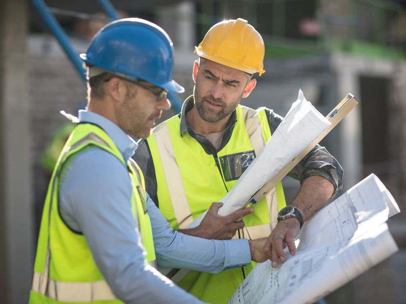 Dos ingenieros discutiendo planos en una obra de construcción, evaluando factores clave para decidir entre alquilar o comprar maquinaria pesada.